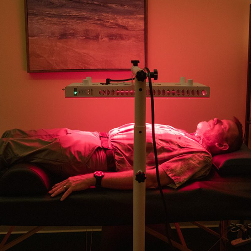 A person lying on a reclined massage bed with red light therapy lamps illuminating the room.