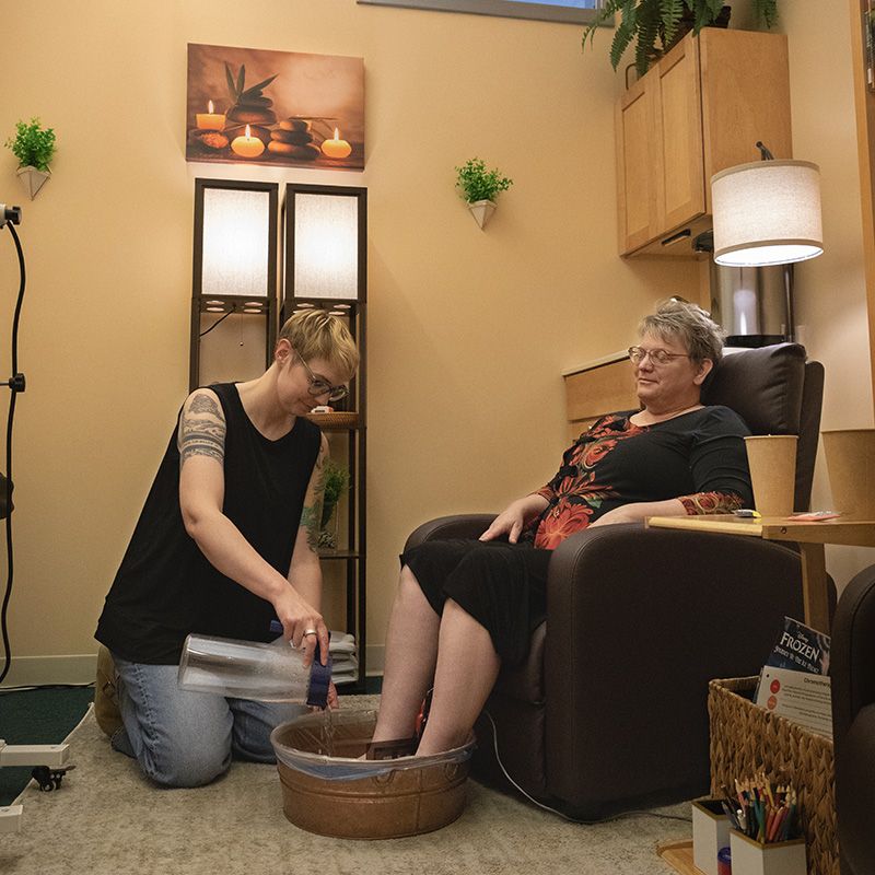 A woman relaxing in a large cushioned chairs receiving a detoxing footbath from a female practitioner.
